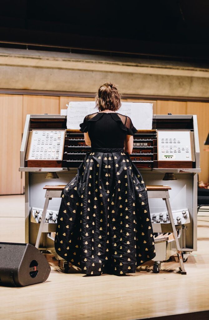 A woman in a patterned dress and black top playing the organ, seen from behind.