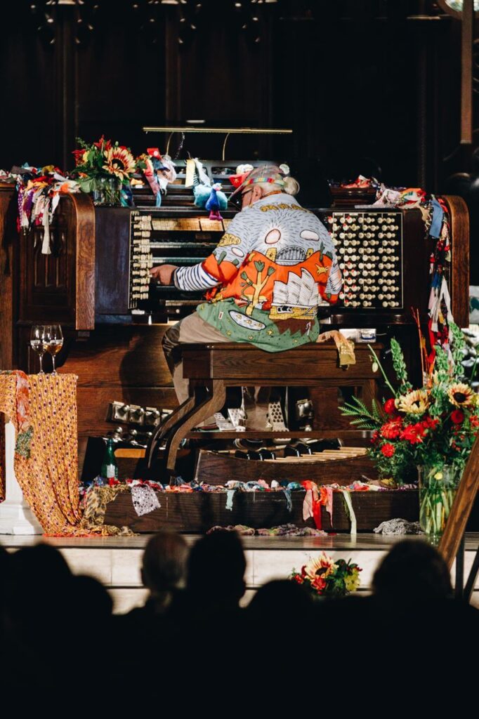 A man in a bright, cartoon-style shirt playing an organ on stage in front of an audience.